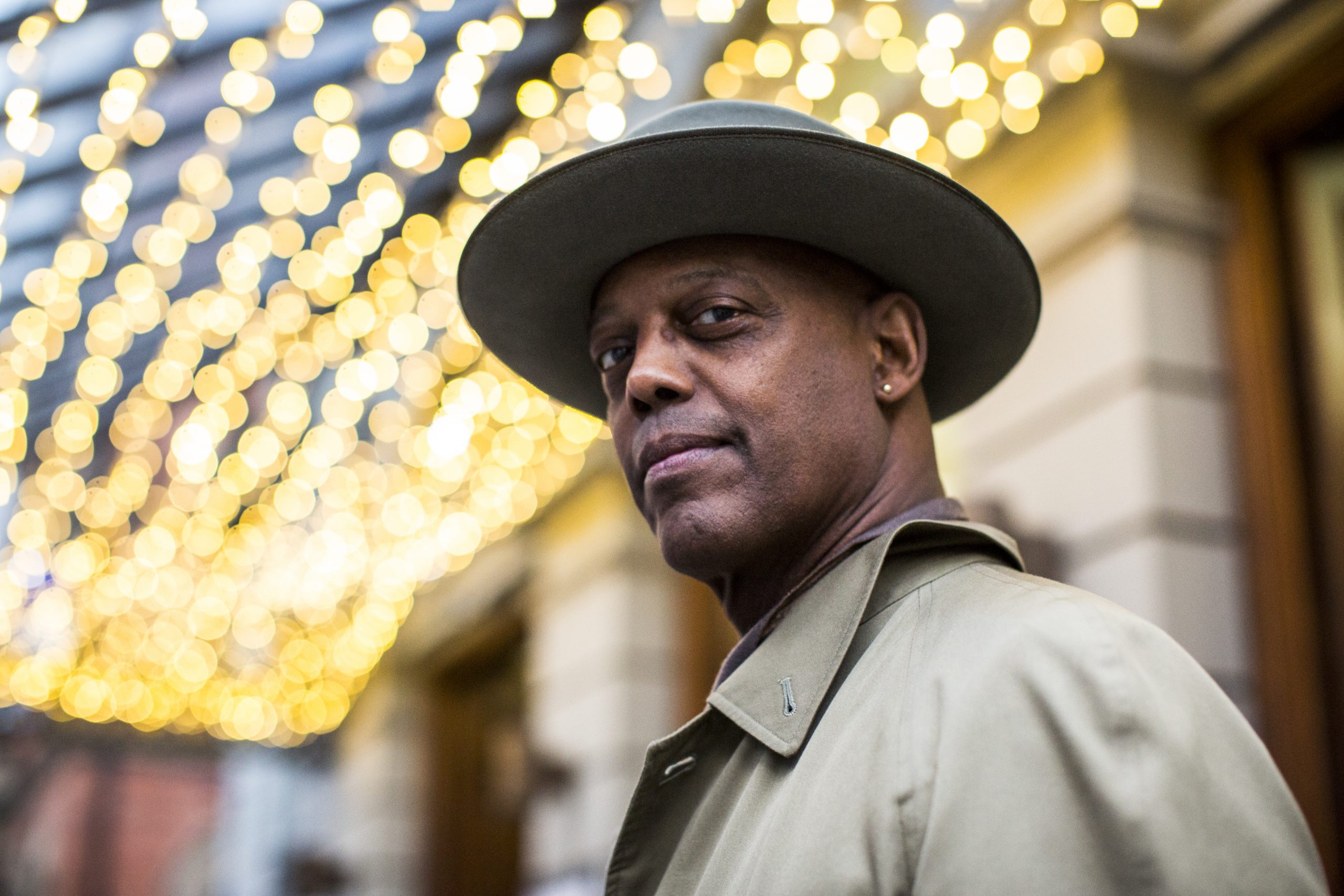Eric Bibb looks looks down at the camera, he is wearing a hat and has fairy lights above him.